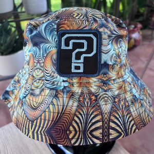 May include: A bucket hat featuring a vibrant, swirling fractal pattern in shades of orange, blue, and yellow. Centered on the front is a rectangular black patch with a light blue embroidered question mark symbol, stitched with visible dark thread.