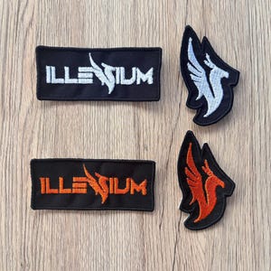 May include: Four embroidered patches. Two rectangular patches with the word "ILLENIUM" in white and orange, and two patches with a stylized bird design in white and orange. All patches are on a black background.