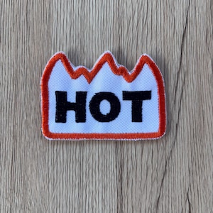 Puede incluir: Un parche blanco con un borde naranja en forma de montaña y un contorno rojo. La palabra "HOT" está impresa en letras negras en negrita en el centro. El parche está sobre una superficie de madera.