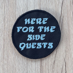 May include: A round, black embroidered patch with the text "HERE FOR THE SIDE QUESTS" in light blue, stylized lettering. The patch has a black border and is set against a light wood grain background.