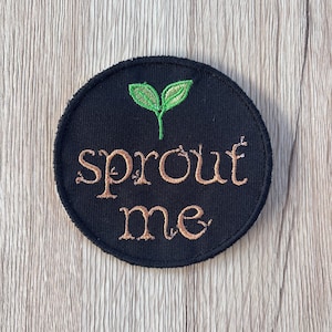 Puede incluir: Parche redondo bordado negro con las palabras "sprout me" en color marrón claro. Un diseño de brote verde está en la parte superior. El parche está sobre una superficie de vetas de madera clara.