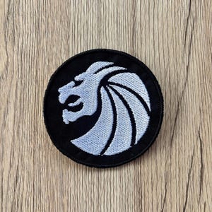 May include: A round, black embroidered patch featuring a white lion's head in profile. The lion has a detailed mane and is set against a solid black background. The patch is likely for clothing or accessories.