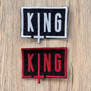 May include: Two rectangular patches with the word "KING" embroidered in white and red, respectively. The patches have a black background and a cross-like symbol below the text. The patches are on a wooden surface.