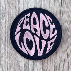 May include: A circular embroidered patch with a black background and light pink stitching. The text PEACE LOVE is embroidered in a stylized, bold font across the center, with a heart shape integrated into the letter O in the word LOVE.