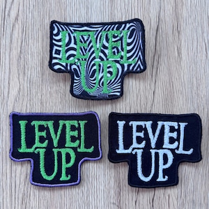 May include: Three embroidered patches shaped like a T-shirt. The top patch has a black and white hypnotic swirl pattern with green text reading LEVEL UP. The bottom left patch is black with green text and a purple border. The bottom right is black with white text.