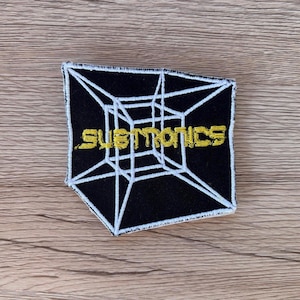 Puede incluir: Parche cuadrado negro con diseño geométrico blanco y texto dorado "SUBTRONICS". El parche tiene un borde blanco y un diseño de cubo 3D. El fondo es de madera.