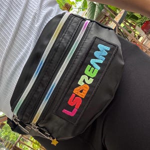 May include: Black fanny pack with rainbow zippers and the word "LSDBEAM" embroidered in colorful letters. The bag has a small gold star charm on the zipper pull. The bag is worn around the waist.