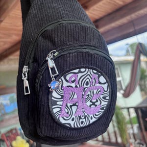 May include: Black corduroy sling bag with a zippered front pocket. The pocket features a circular patch with a black and white swirl design and the embroidered letters "FB" in purple. A small charm hangs from the zipper.