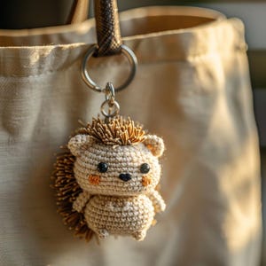 Peut inclure: Un porte-clés en forme de hérisson crocheté. Le hérisson a un corps brun clair, des yeux foncés et des joues orangées. Il est doté d'un dos épineux et d'un petit nez noir. Le porte-clés est accroché à un sac fourre-tout beige.