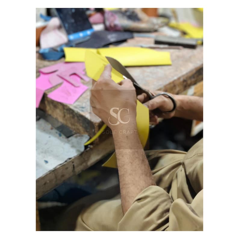 May include: A person using scissors to cut a piece of yellow material. The image shows a close-up of the hands and the material being cut. The logo "SC SOLE CRAFT" is visible on the person's arm. Other pieces of material are visible in the background.