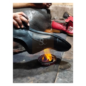 May include: A shoemaker working on a black leather shoe, using a flame to shape the sole. The image shows the process of shoe construction, with a red shoe in the background. The text "SOLE CRAFT" is visible on the shoe.