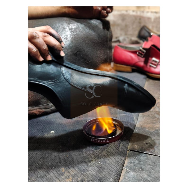 May include: A shoemaker working on a black leather shoe, using a flame to shape the sole. The image shows the process of shoe construction, with a red shoe in the background. The text "SOLE CRAFT" is visible on the shoe.