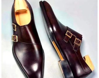 May include: A pair of burgundy leather double monk strap shoes. The shoes have a polished finish, gold-colored buckles, and a pointed toe. Wooden shoe trees are inside the shoes.