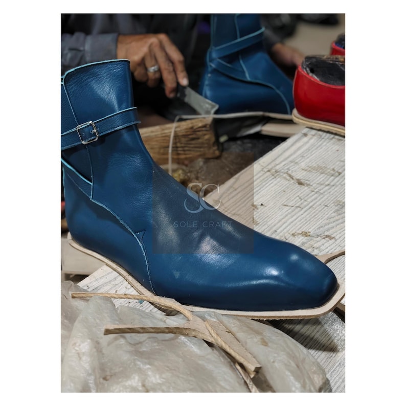 May include: A close-up of a blue leather boot with a buckle strap. The boot has a square toe and is being crafted on a wooden surface. The image also shows a partially visible red boot and the text "SOLE CRAFT".