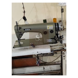 May include: A Brother industrial sewing machine in a workshop setting. The machine is a muted green color and sits on a white table. Spools of thread in yellow and green are visible. The machine has a light attached and the Brother logo is visible.