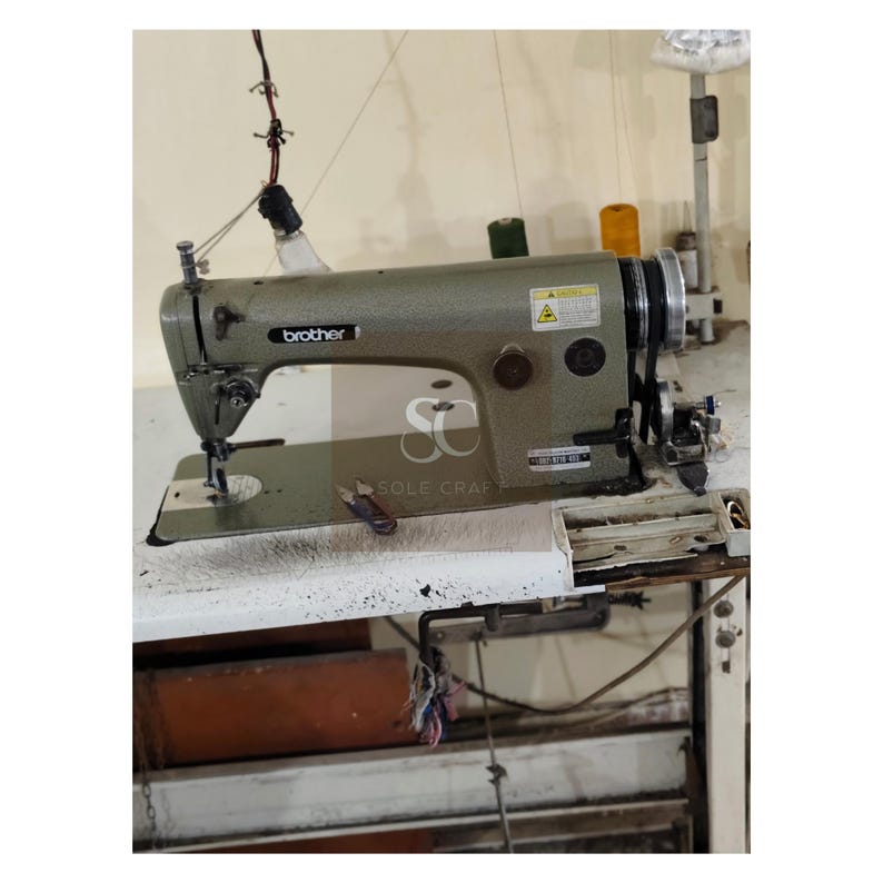 May include: A Brother industrial sewing machine in a workshop setting. The machine is a muted green color and sits on a white table. Spools of thread in yellow and green are visible. The machine has a light attached and the Brother logo is visible.