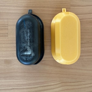 May include: Two oblong containers, one black and one yellow, sit side-by-side on a wooden surface. The black container has a textured surface and a small handle. The yellow container is smooth and has a small rectangular handle.