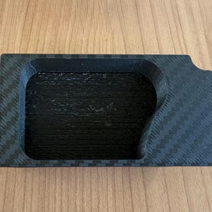 Puede incluir: Una bandeja negra con un patrón texturizado de fibra de carbono. La bandeja tiene una forma rectangular con una hendidura curva en un lado.