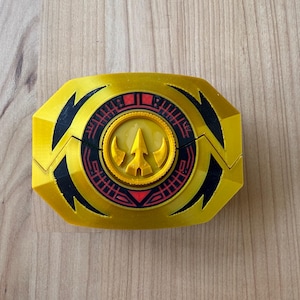 Puede incluir: Una hebilla de cinturón Power Rangers amarilla y negra con un emblema circular en el centro. El emblema tiene un diseño rojo y negro con un símbolo amarillo en forma de flecha en el medio. La hebilla es de plástico.