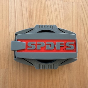 Pode incluir: Um objeto retangular cinza e vermelho com as letras "SPDFS" em cinza. O objeto tem uma abertura circular e está sobre uma superfície de madeira.