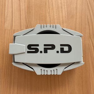 Puede incluir: Una insignia rectangular de color gris claro con una cubierta central abatible que presenta el texto negro S.P.D. La insignia tiene un acabado texturizado, una pequeña pestaña en el lado izquierdo y detalles ranurados decorativos en los bordes superior e inferior.