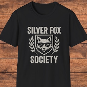 Könnte beinhalten: Schwarzes T-Shirt mit dem Text "SILVER FOX SOCIETY" in verblichener, weißer Schrift. Das Design zeigt einen Fuchskopf mit Sonnenbrille in einem Schild, umgeben von Lorbeerblättern.