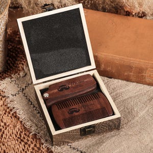 Könnte beinhalten: Ein Bartpflege-Set aus Holz in einer hellen Holzkiste. Die Kiste enthält einen Kamm und eine Bürste, beide mit einem Schnurrbart-Motiv und dem Wort "Hmolen". Die Box ist geöffnet und steht auf einer neutralfarbenen Oberfläche.