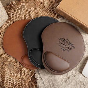 Könnte beinhalten: Drei Leder-Mauspads in Braun, Schwarz und Beige, jeweils mit Handgelenkauflage. Das braune Pad ist mit dem Namen "KEEGAN" und dekorativen Verzierungen personalisiert. Eine weiße kabellose Maus und ein Buch sind ebenfalls zu sehen.