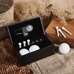 May include: A black leather golf kit with a divot repair tool, golf tees, and two golf balls. Three additional golf tees and a golf ball are placed outside the kit. The kit is on a textured surface.