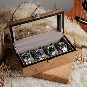 May include: A wooden watch display case with a glass top, holding four wristwatches with silver and gold bands. The watches have black, blue, and green faces. The case is open, revealing a gray interior. A woven basket and ukulele are in the background.