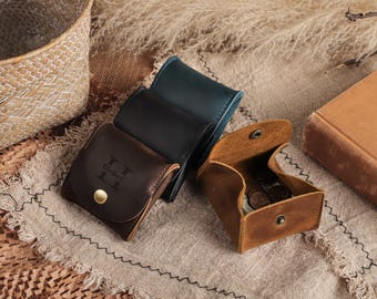May include: A collection of leather coin purses in brown, black, and teal. One purse is open, revealing coins inside. The brown purse has a gold snap closure and an embossed initial. The purses are on a neutral-toned fabric.