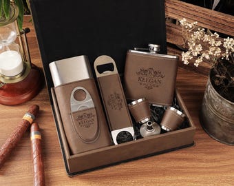 May include: A brown leatherette gift set including a cigar cutter, bottle opener, flask, shot glasses, and a box. The items are engraved with the name "Keegan". Two cigars are on the wooden surface.