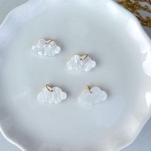 May include: Four cloud-shaped earrings with gold-colored hoops. The earrings are white with a marbled effect, resting on a white, scalloped plate. The earrings are small, delicate, and suitable for everyday wear.
