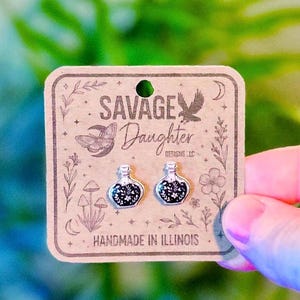 May include: A pair of silver-toned potion bottle stud earrings with black and white floral details, displayed on a square brown cardboard backing card printed with the text SAVAGE Daughter DESIGNS LLC and HANDMADE IN ILLINOIS.