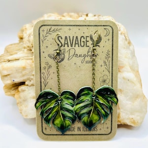 May include: A pair of dangle earrings featuring a green monstera leaf design. The earrings are attached to a gold-colored chain and hook. The earrings are displayed on a brown Savage Daughter Designs LLC card. The card reads "MADE IN ILLINOIS".