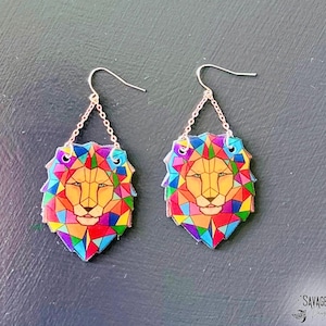 May include: A pair of colorful lion head earrings. The earrings feature a geometric design with a yellow face and a rainbow mane. The earrings are suspended from silver-colored ear wires and chains.