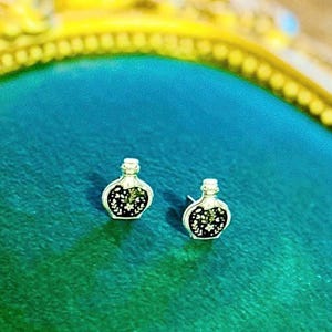 May include: A pair of silver-toned stud earrings shaped like small potion bottles. Each earring features a black center inlay decorated with a white floral and leaf pattern, finished with a metallic silver border and a standard post backing.