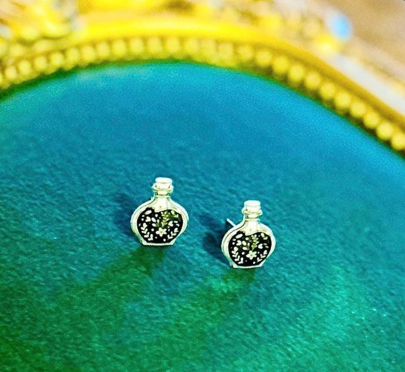 May include: A pair of silver-toned stud earrings shaped like small potion bottles. Each earring features a black center inlay decorated with a delicate white floral and leaf pattern, secured with a standard post backing for earlobe attachment.