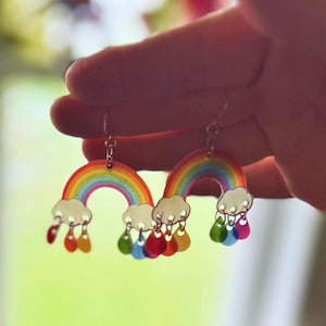 May include: A pair of rainbow-themed earrings. Each earring features a colorful rainbow arch with white cloud accents and dangling teardrops in various colors. The earrings are held up by a silver chain.