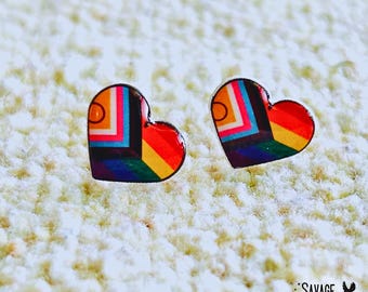 May include: A pair of heart-shaped earrings with a colorful geometric design. The earrings feature a black outline and a pattern of orange, red, yellow, blue, and green. The earrings are on a textured, off-white surface.