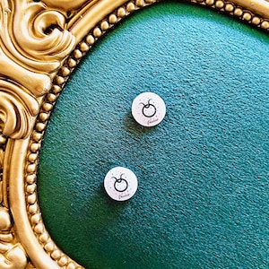 May include: Two white Taurus zodiac sign magnets with black illustrations and text, set against a teal textured background. The magnets are round and have a black border. The background is framed by a gold ornate border.
