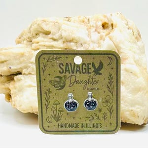 May include: Silver-toned stud earrings featuring a potion bottle design with a dark blue and white starry pattern. The earrings are displayed on a card with the text "SAVAGE DAUGHTER DESIGNS LLC" and "HANDMADE IN ILLINOIS".