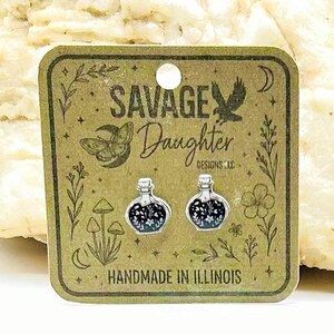 May include: A pair of silver-toned potion bottle stud earrings featuring a black enamel fill with white floral details. The earrings are displayed on a square brown card printed with the text SAVAGE Daughter DESIGNS LLC and HANDMADE IN ILLINOIS.