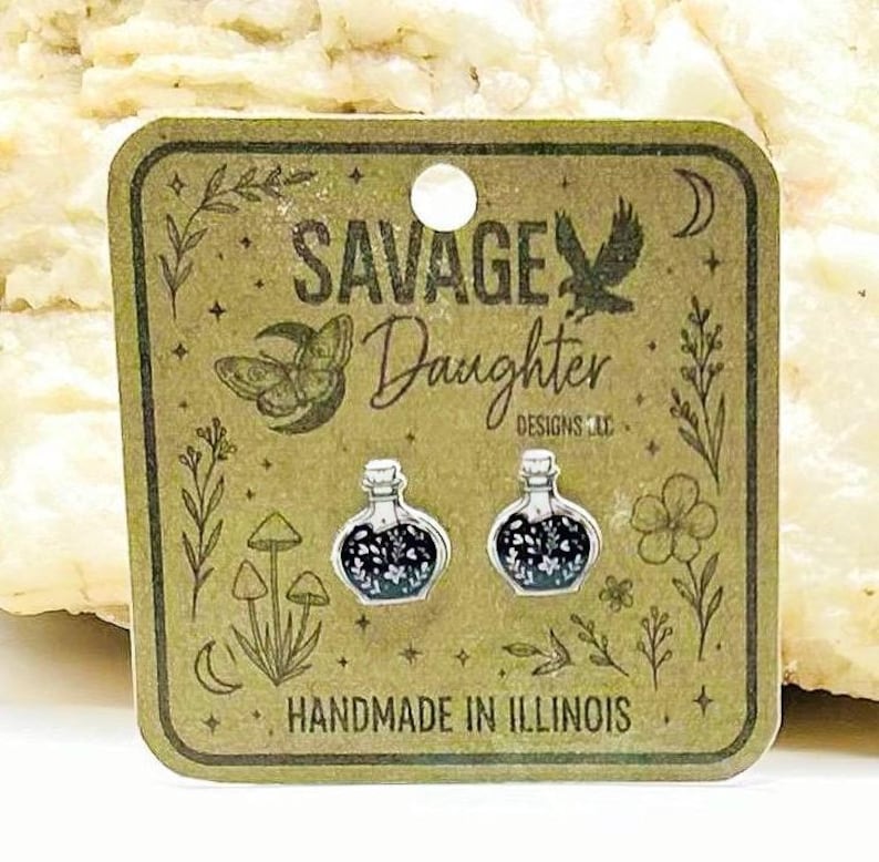 May include: A pair of silver-toned potion bottle stud earrings featuring a black enamel fill with white floral details. The earrings are displayed on a square brown card printed with the text SAVAGE Daughter DESIGNS LLC and HANDMADE IN ILLINOIS.