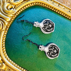 May include: A pair of black and white potion bottle earrings with floral designs. The earrings hang from black chains and hooks. The earrings are displayed on a green surface, framed by a gold-colored ornate border.