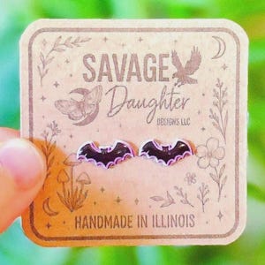 May include: A pair of black bat-shaped stud earrings with purple outlines, displayed on a square tan cardboard backing card. The card features the text SAVAGE Daughter DESIGNS LLC, a moth illustration, floral motifs, and HANDMADE IN ILLINOIS.