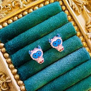 May include: Two small, colorful yeti-shaped earrings with blue eyes and horns rest on a teal velvet jewelry display. The earrings have a white outline and a pink mouth. The display is in a gold-colored ornate frame.