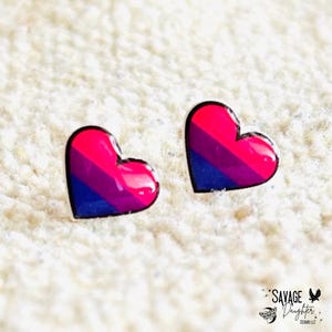 May include: Two heart-shaped earrings with a glossy finish. Each earring features a design with pink, purple, and blue stripes, outlined in black. The earrings are set against a textured white background.