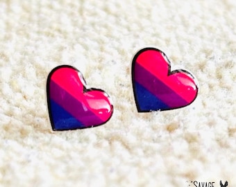 May include: Two heart-shaped earrings with a glossy finish. Each earring features a design with pink, purple, and blue stripes, outlined in black. The earrings are set against a textured white background.