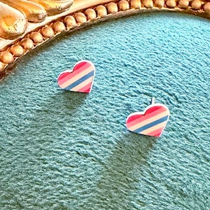 May include: A pair of heart-shaped stud earrings. Each earring features a pink, white, and blue striped design. The earrings are set against a teal background, with a gold-colored decorative frame in the upper left corner.